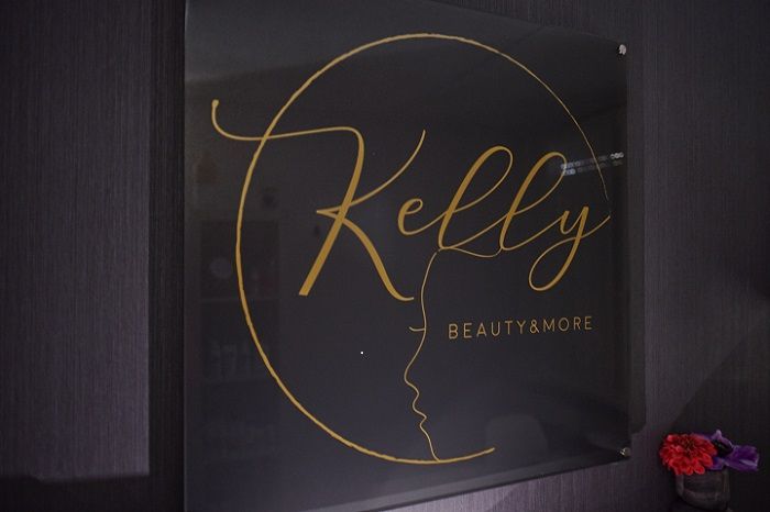 Het logo van Kelly Beauty&More in goud op de donkere wand van de salon