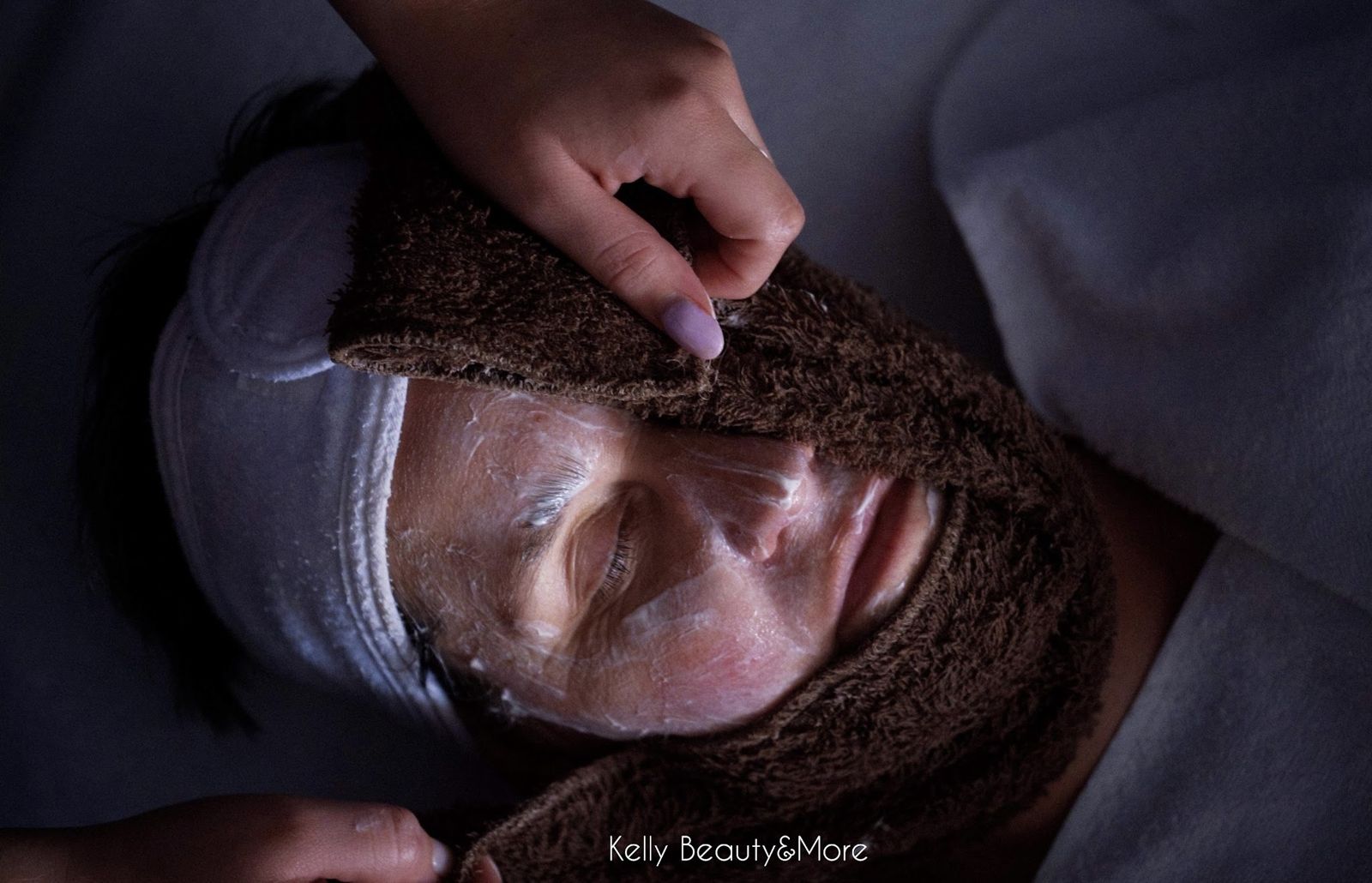 Cliënte met haarband en masker krijgt een warme doek tijdens een gezichtsbehandeling